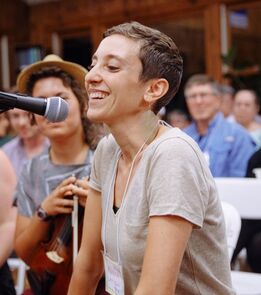 A headshot of Rabbi Mónica Gomery, a light-skinned woman with short hair and a gray shirt, singing into a microphone.