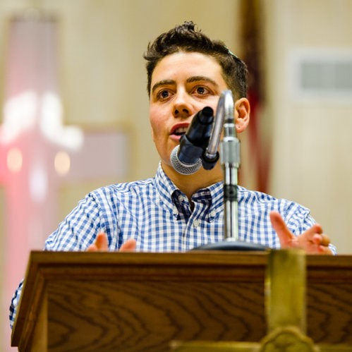 A headshot of Rabbi Ari Lev Fornari, a light-skinned man with short brown hair and a blue checkered shirt, giving a drash behind a microphone at a podium.