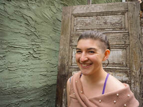 A headshot of Miryam Coppersmith, a light-skinned woman smiling in front of an antique door and textured stucco wall.